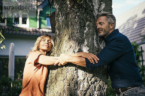 Lächelnde reife Männer und Frauen umarmen an einem sonnigen Tag einen Baum im Hinterhof