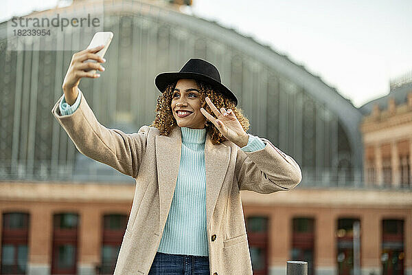 Glückliche Frau mit Hut gestikuliert und macht ein Selfie mit dem Handy