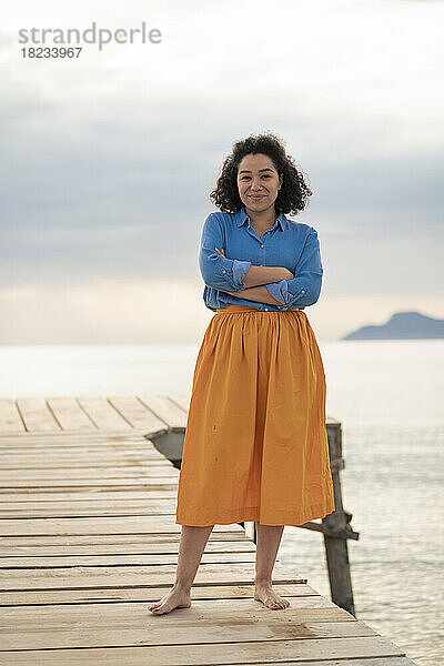 Confident woman with arms crossed standing on jetty at sunset