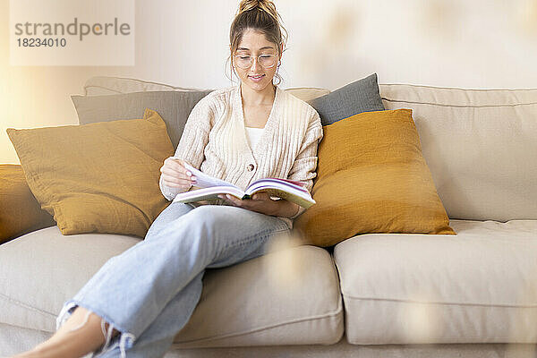 Lächelnde junge Frau liest zu Hause auf dem Sofa ein Buch