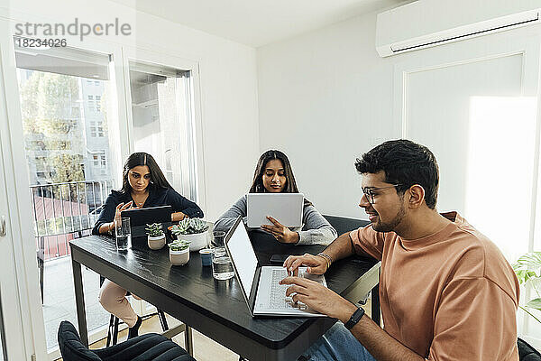 Lächelnder Mann mit Laptop vor Frauen  die über Tablet-PCs auf dem Tisch im Wohnzimmer lernen
