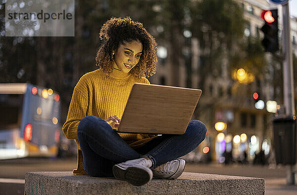 Lächelnde Geschäftsfrau benutzt Laptop in der Abenddämmerung auf Beton