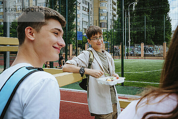 Lächelnder Junge mit Brotdose steht mit Freunden auf dem Schulhof