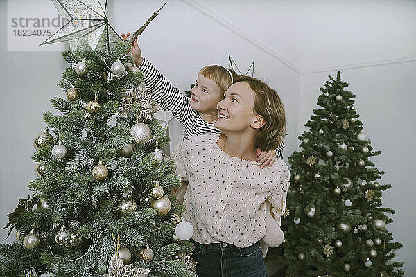 Lächelnde Mutter gibt ihrer Tochter eine Huckepackfahrt  um einen Stern auf den Weihnachtsbaum zu setzen