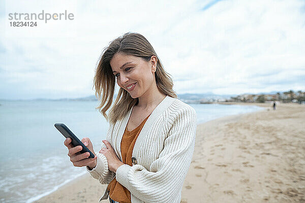 Lächelnde schöne junge Frau  die am Strand ihr Smartphone benutzt