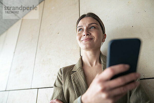 Lächelnde Geschäftsfrau mit Mobiltelefon vor der Wand