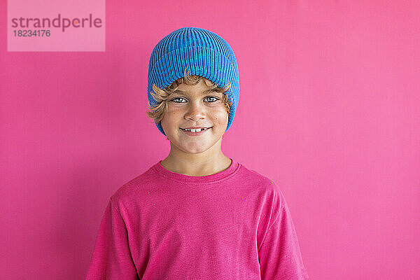 Lächelnder Junge mit blauer Strickmütze vor rosa Hintergrund