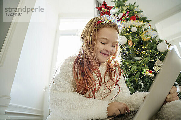 Glückliches Mädchen mit Laptop  das zu Hause vor dem Weihnachtsbaum sitzt