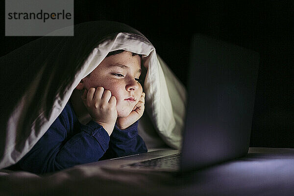 Junge ist kurz davor einzuschlafen und schaut im Schlafzimmer auf den Laptop