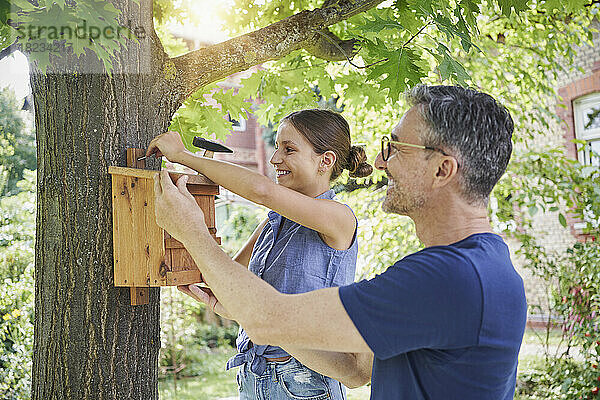 Glücklicher Mann mit Tochter hängt Nistkasten an Baumstamm im Garten