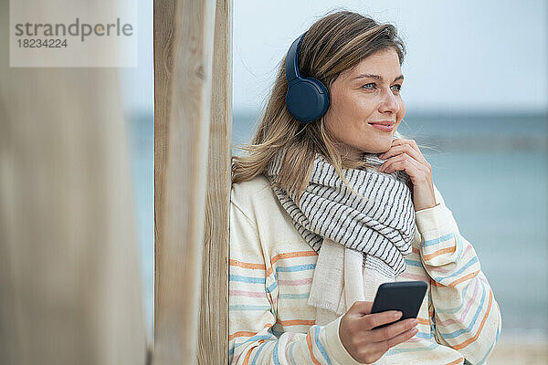 Lächelnde schöne junge Frau hält Smartphone in der Hand und hört Musik am Strand