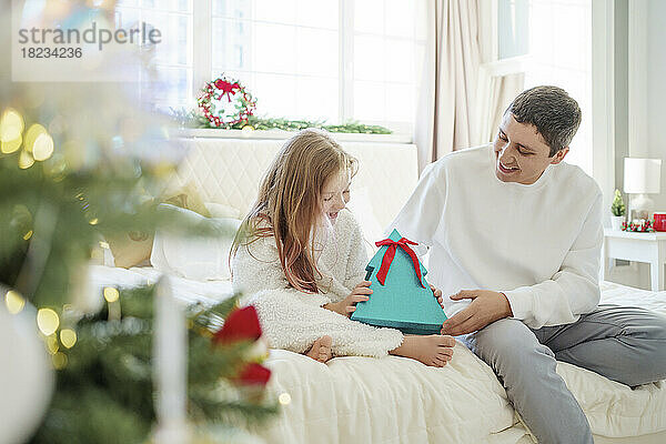 Glückliches Mädchen hält Weihnachtsgeschenk in der Hand  während der Vater zu Hause auf dem Bett sitzt