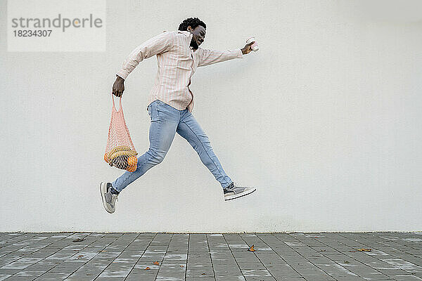 Mann mit Einkaufstüte und Einwegkaffeetasse springt vor weiße Wand