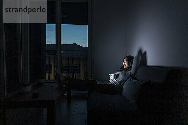 Lächelnde Frau mit Kaffeetasse  die zu Hause einen Film auf dem Laptop anschaut