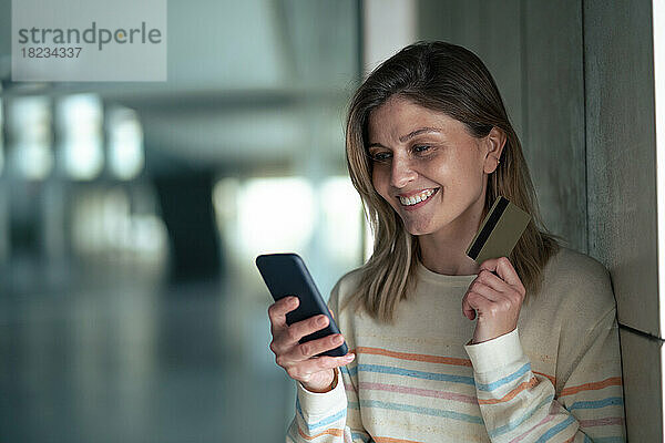 Lächelnde junge Frau hält Smartphone und Kreditkarte in der Hand und lehnt an der Wand