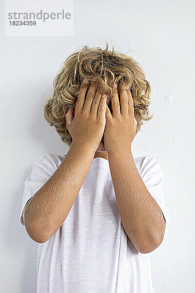 Schüchterner Junge bedeckt Gesicht mit Händen vor weißem Hintergrund