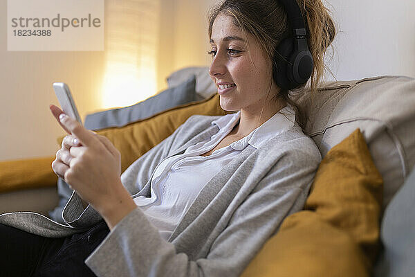 Lächelnde Frau mit kabellosen Kopfhörern und Smartphone auf dem heimischen Sofa