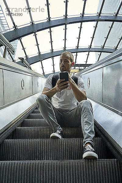 Mann sitzt mit Smartphone auf Rolltreppe