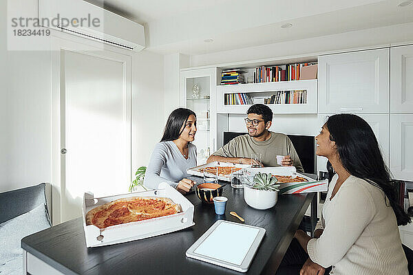 Glückliche Frauen und Männer diskutieren und essen Pizza am Esstisch im Wohnzimmer