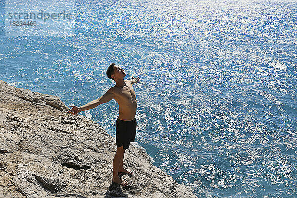 Carefree man with arms outstretched standing on rock by sea