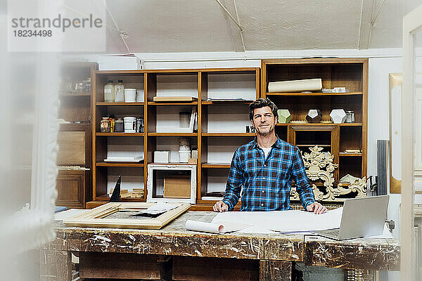 Lächelnder Handwerker steht an der Werkbank in der Werkstatt