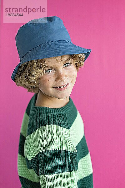 Lächelnder Junge mit Fischerhut und Pullover vor rosa Hintergrund