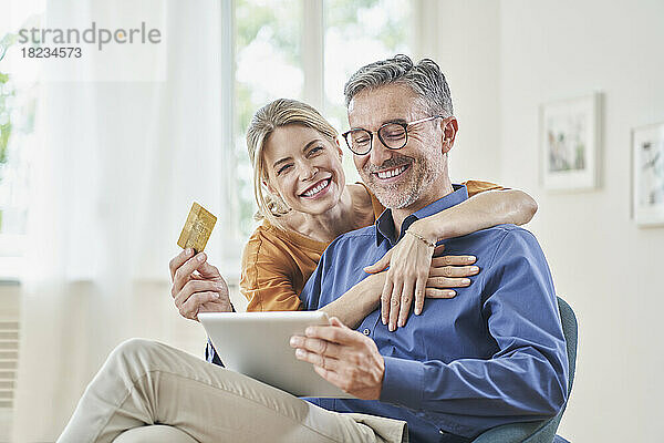 Glückliche Frau umarmt Mann mit Kreditkarte beim Online-Shopping per Tablet-PC zu Hause