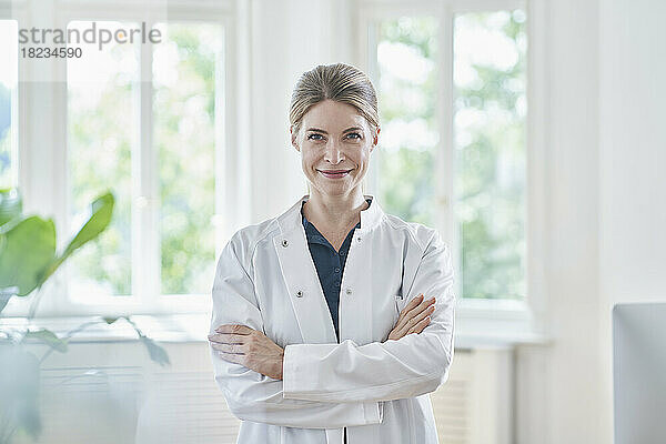 Smiling female doctor in lab coat with arms crossed at medical practice