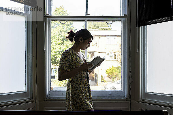 Lächelnde Frau liest ein Buch und steht vor dem Fenster