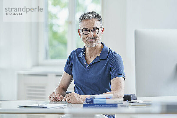 Confident doctor wearing eyeglasses sitting at desk in medical practice