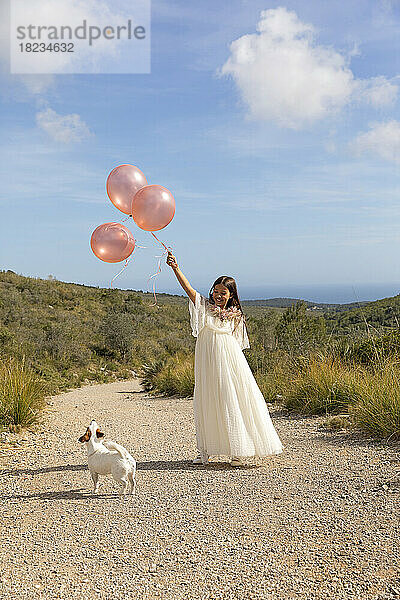 Verspieltes Mädchen in weißem Kleid hält Luftballons mit Hund auf dem Weg