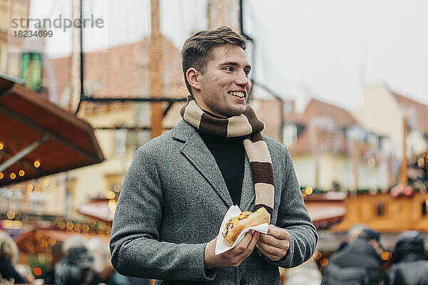 Glücklicher junger Mann mit Hot Dog steht auf dem Weihnachtsmarkt