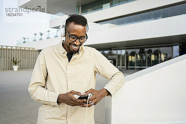 Glücklicher Mann benutzt Smartphone und trägt Kopfhörer vor dem Gebäude