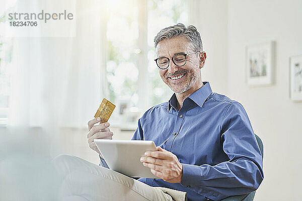 Glücklicher Mann mit Kreditkarte  der zu Hause über einen Tablet-PC Online-Einkäufe tätigt