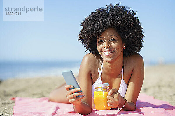 Lächelnde junge Frau entspannt sich an einem sonnigen Tag mit Smartphone und Orangensaft am Strand