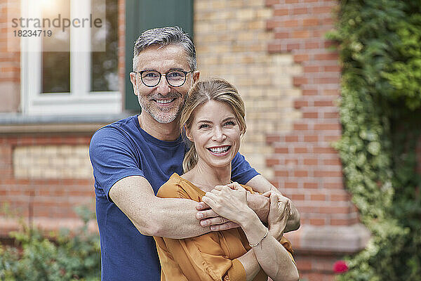 Lächelnder Mann umarmt Frau von hinten vor dem Haus