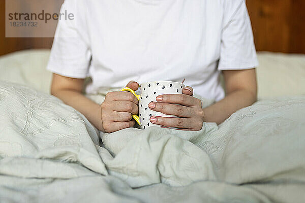 Hände einer jungen Frau mit Kaffeetasse  die zu Hause auf dem Bett sitzt