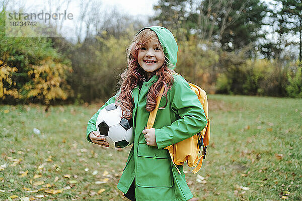 Glückliches Mädchen steht mit Rucksack und Fußball im Park