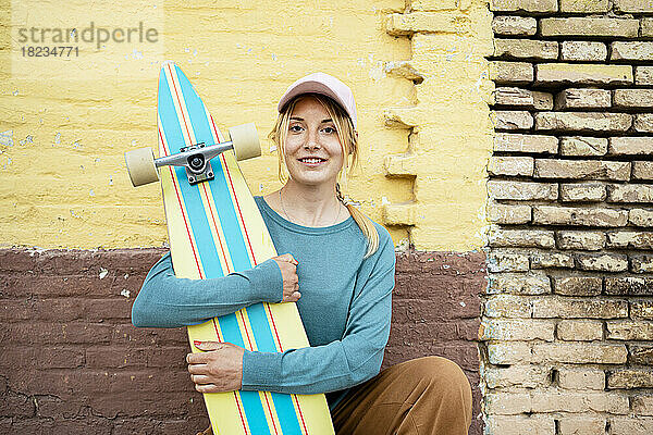 Lächelnde Frau mit Mütze kauert mit Skateboard vor der Wand