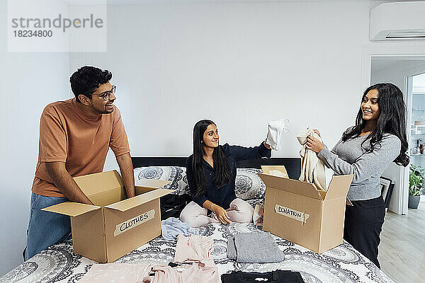 Glückliche Frauen und Männer packen zu Hause Kisten mit Kleidung für eine Spende auf dem Bett