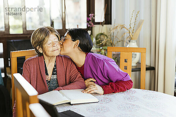 Pflegekraft küsst ältere Frau  die zu Hause am Tisch sitzt
