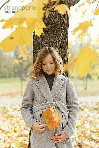 Schwangere Frau mit Ahornblatt lehnt an Baum im Herbstpark