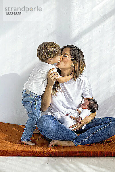 Sohn küsst Mutter  die mit neugeborenem Mädchen zu Hause sitzt