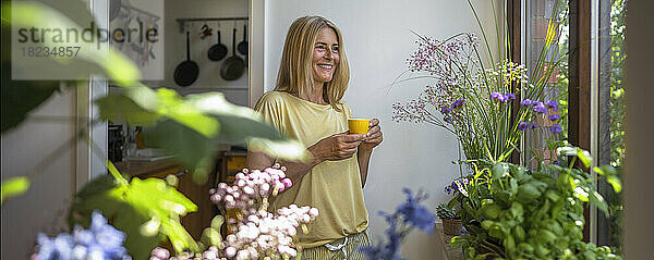 Glückliche reife Frau hält eine Tasse Kaffee in der Hand und steht zu Hause am Fenster
