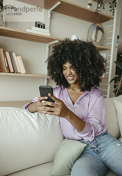 Lächelnde Afro-Frau benutzt Smartphone und sitzt zu Hause auf dem Sofa