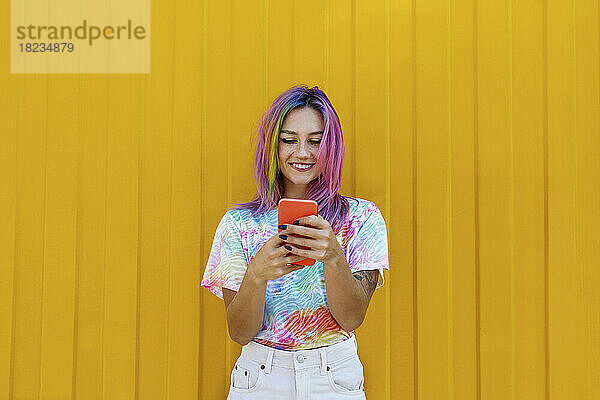 Lächelnde junge Frau im Batik-T-Shirt mit Smartphone vor gelber Wand