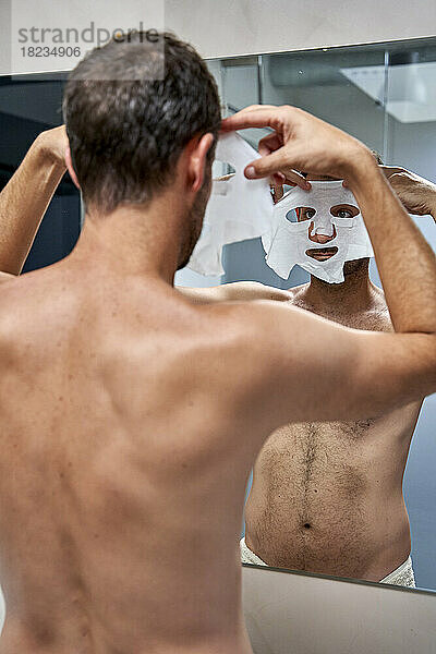 Mann trägt zu Hause vor dem Spiegel eine Blattmaske auf sein Gesicht auf