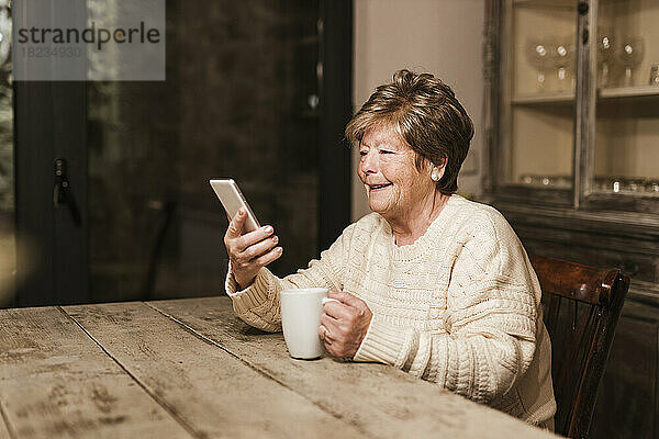 Lächelnde ältere Frau benutzt Smartphone und sitzt mit einer Kaffeetasse zu Hause