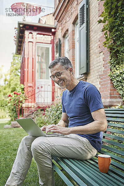 Glücklicher Geschäftsmann sitzt mit Laptop auf Bank im Hinterhof
