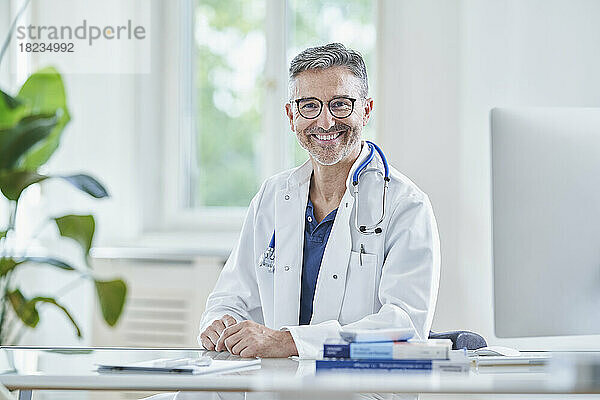Happy mature doctor in lab coat sitting in medical practice